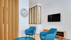Una habitación moderna con una mesa redonda de vidrio, dos sillones azules, un armario de madera, un televisor montado en la pared y un reloj grande.
