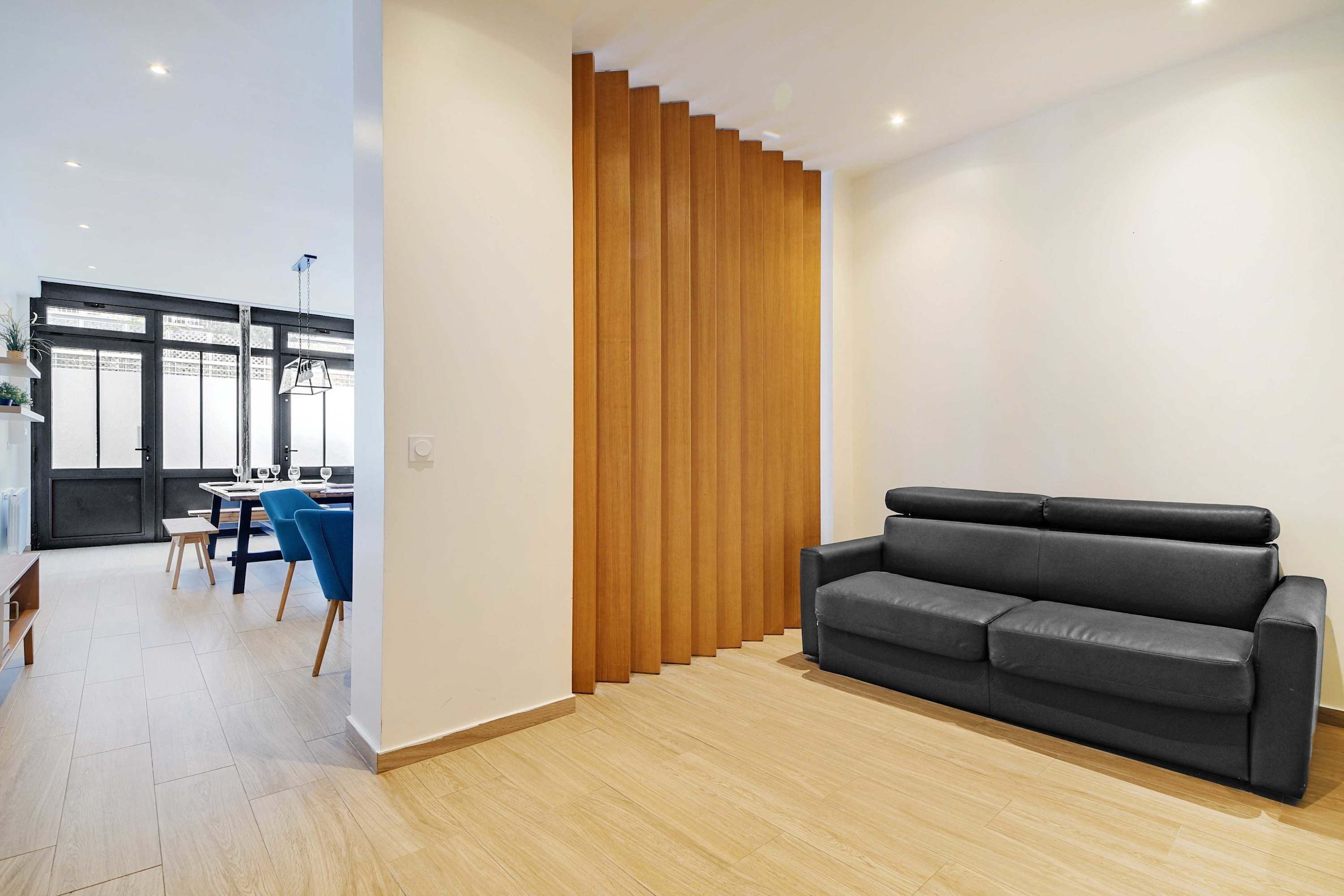 A modern living room with a black leather sofa, wooden slatted wall, and a dining area with blue chairs.