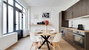 A modern kitchen with a round dining table, white chairs, a built-in oven, and a large window providing natural light. - Amazing Apartment - 2BR 6P - Bastille (Paris)