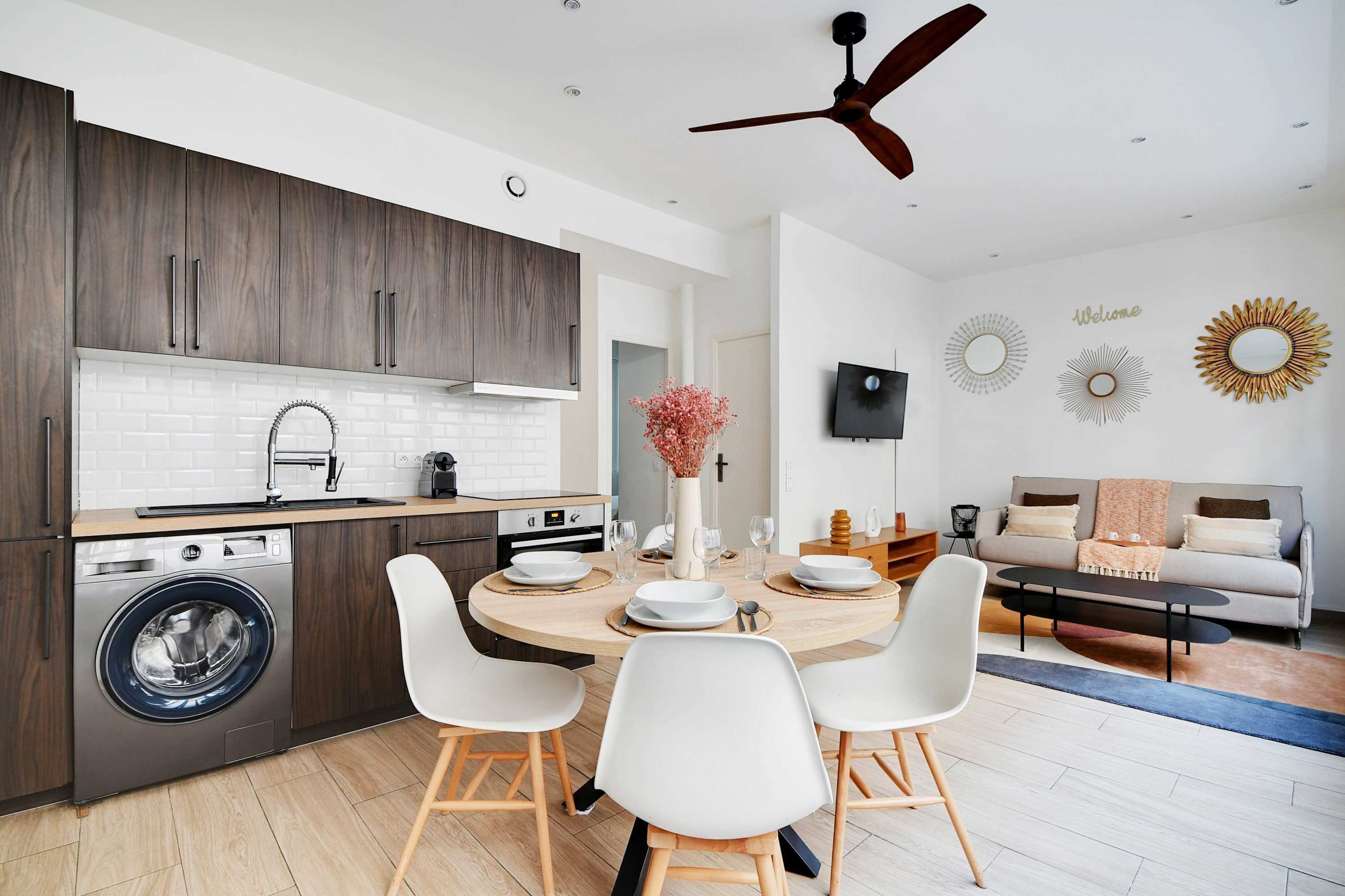 A modern kitchen with a built-in washing machine, a dining area with a round table and white chairs, and a living area with a sofa and decorative wall art.