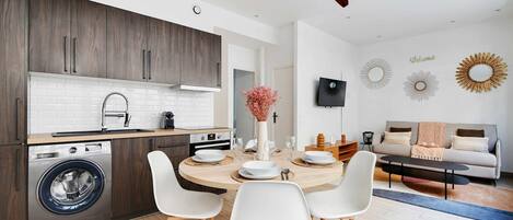 A modern kitchen with a built-in washing machine, a dining area with a round table and white chairs, and a living area with a sofa and decorative wall art.