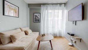 Un salon moderne avec un canapé beige, une table basse ronde en bois, un téléviseur à écran plat fixé au mur et une grande fenêtre ornée de rideaux légers.