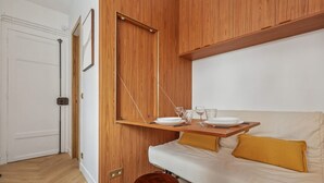 Une pièce avec un mur recouvert de panneaux de bois, un canapé blanc agrémenté de coussins couleur moutarde, un tabouret en bois et une petite table sur laquelle sont disposées des assiettes et des verres.