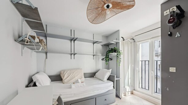 A modern bedroom with a bed, built-in shelves, a hanging light fixture, and a large window. - Cozy Studio - Rue Saint Maur - Mobility Lease (Paris)