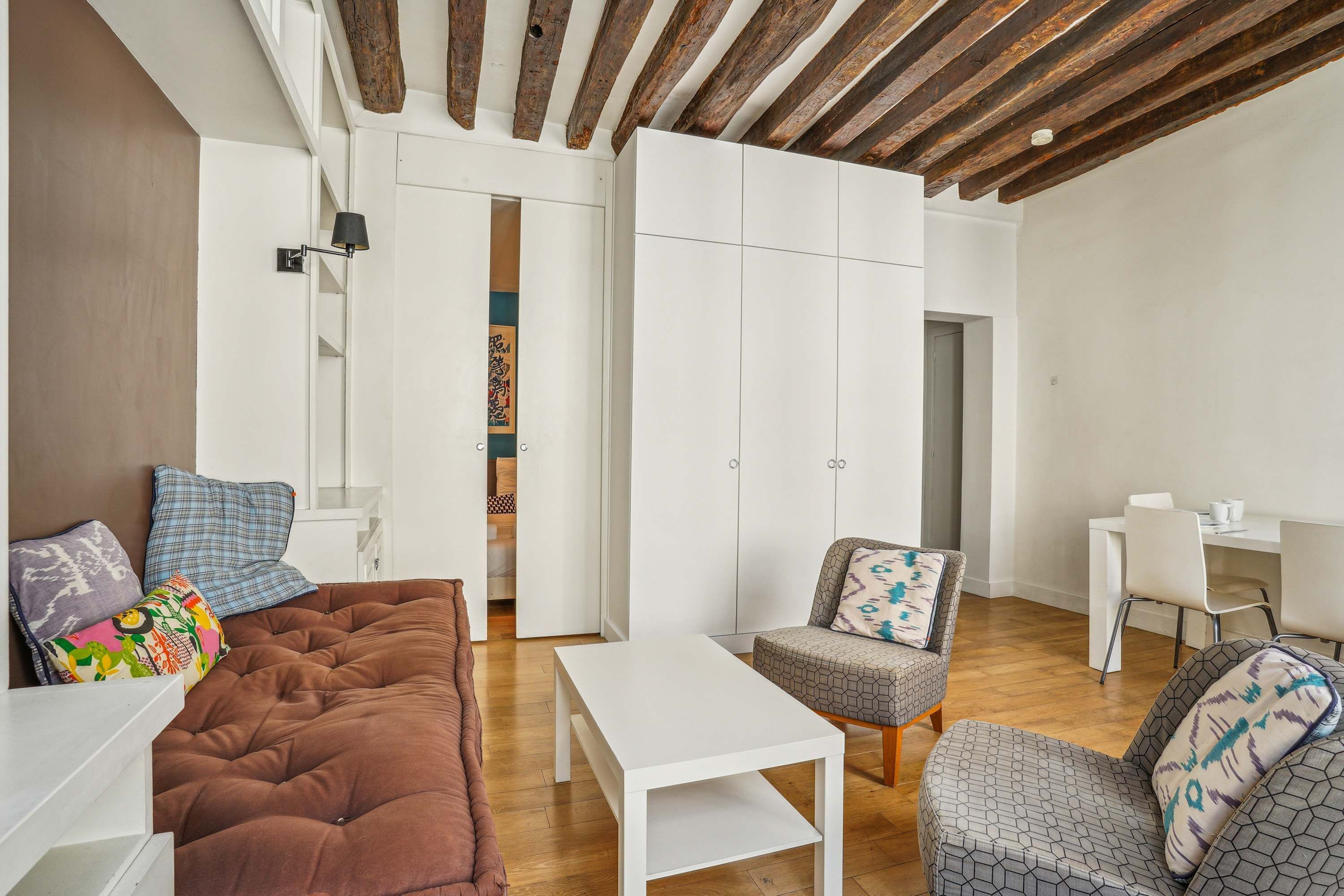 A modern living room with a brown sofa, a white table, and patterned chairs. There is a wardrobe with white doors and a door with artwork.