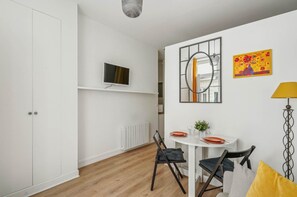 A modern room with a white wardrobe, a TV mounted on a shelf, a dining table with chairs, and a mirror on the wall. - Cosy 2P Studio in the Heart of the Marais (Paris)