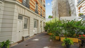 Une ruelle étroite entre deux bâtiments, l’un avec une façade blanche et l’autre en briques, doté de balcons. Des plantes en pot bordent la ruelle.