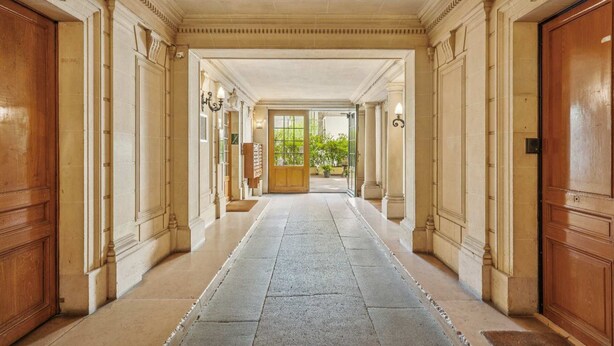 Un vaste hall doté d’un sol en pierre, bordé de portes et de colonnes en bois, qui mène à une grande porte en bois au fond.