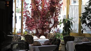 A restaurant with tables set for dining, surrounded by potted plants and a large tree with red leaves.