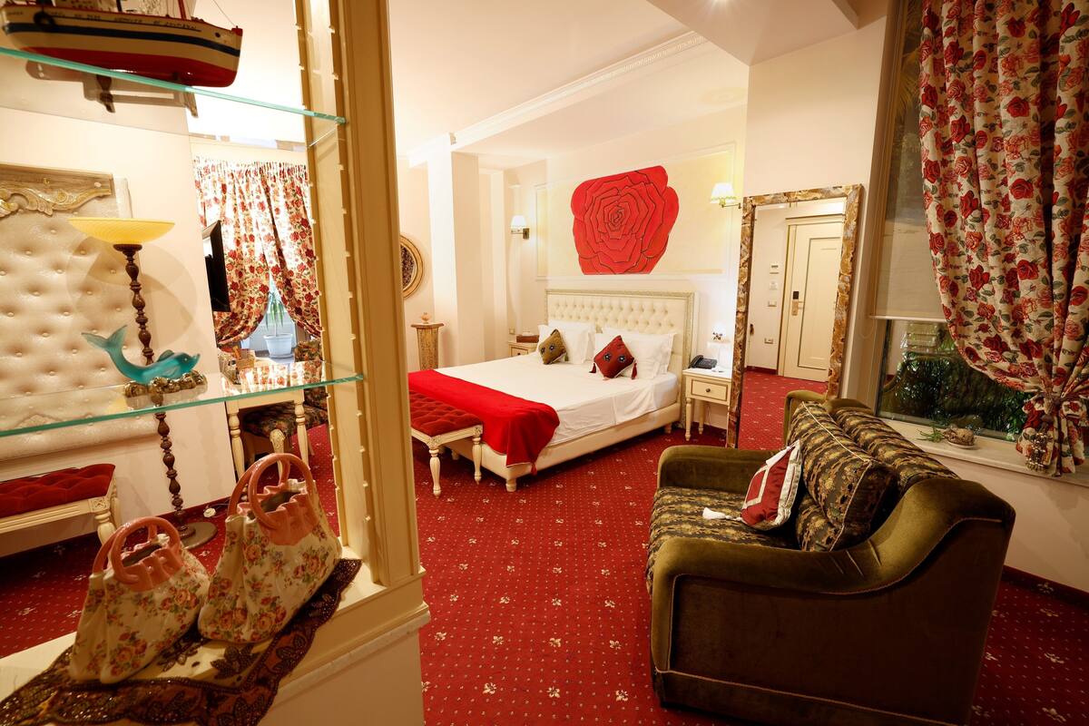 Ein Schlafzimmer mit einem großen Bett, einem grünen Sofa, einer Vitrine mit Handtaschen und einem roten, floralen Vorhang.