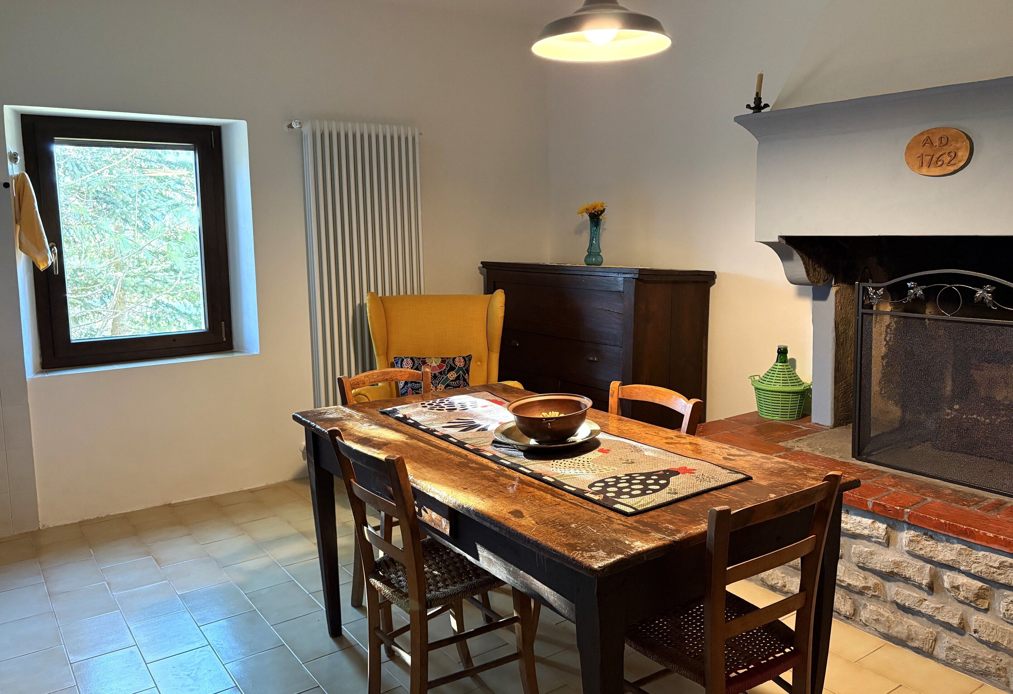 A dining area with a wooden table, chairs, a window with blinds, a radiator, a fireplace, and a wall-mounted sign.