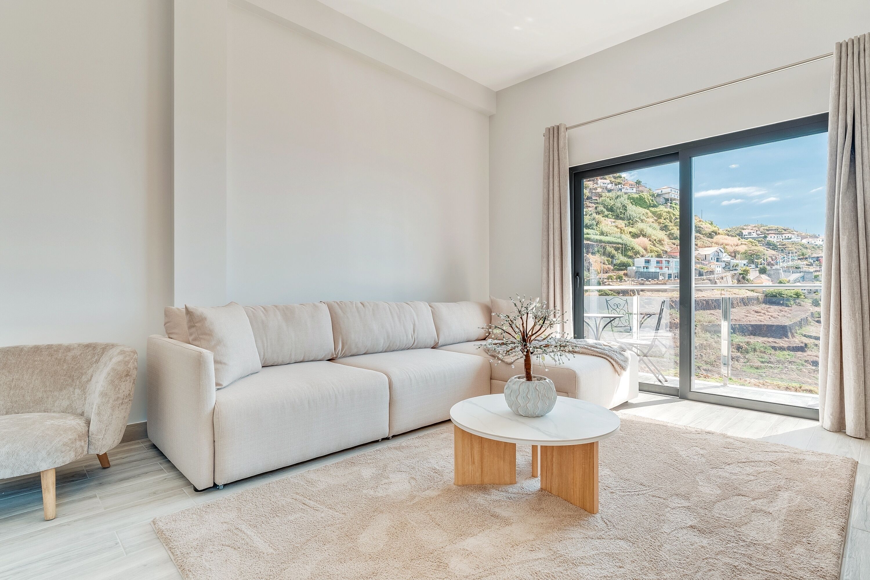 Una sala de estar moderna con un sofá color beige, una mesa de centro redonda y vistas a un pueblo en la ladera a través de grandes ventanales.