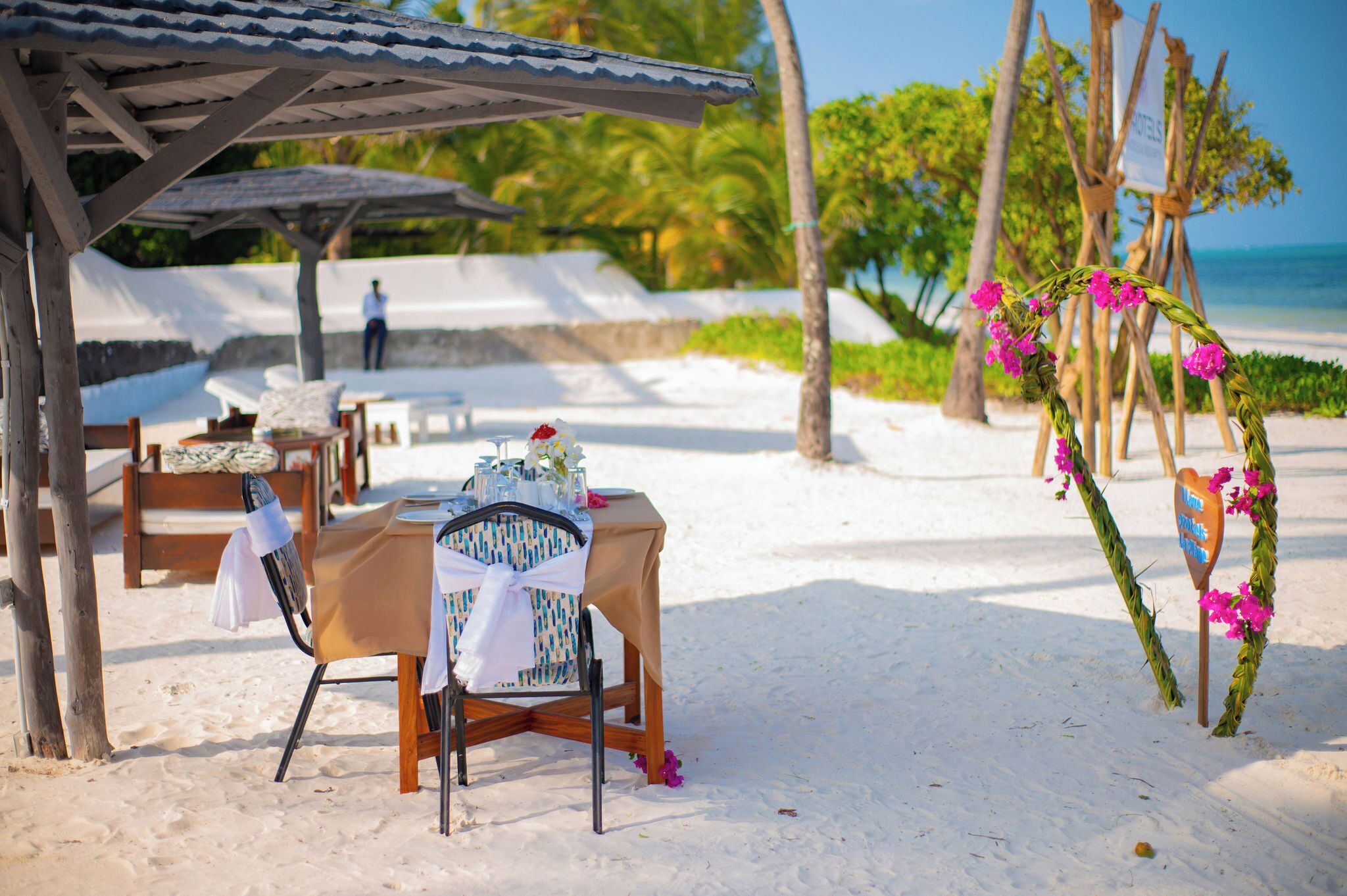 A beachside setting with a table set for a meal, a wooden chair, and a decorative archway with flowers.