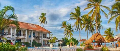 A beachfront hotel with a pool, palm trees, and a thatched-roof structure.