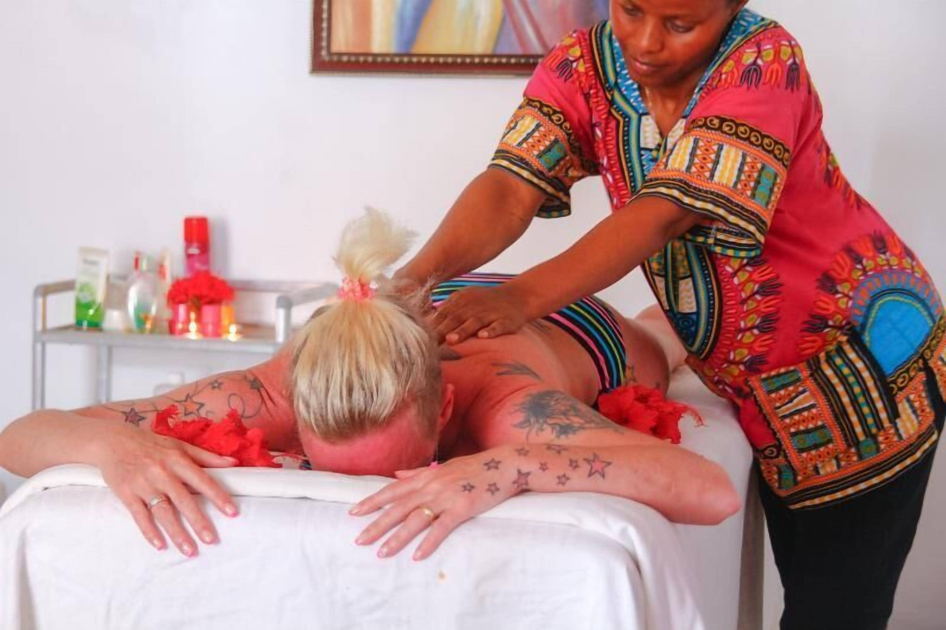 A person receiving a massage in a room with a white bed and a small table with various bottles and a red flower.