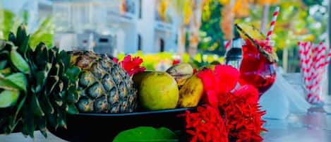 Una mesa con una cesta de frutas que incluye piña, guayaba y otras frutas, junto con flores rojas y una bebida con pajita.