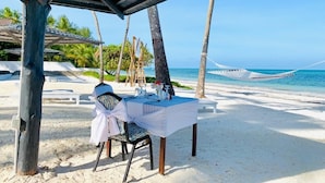 A beachside table set with a white tablecloth, a chair, and a hammock in the background. - Protels La Plage (Bwejuu)