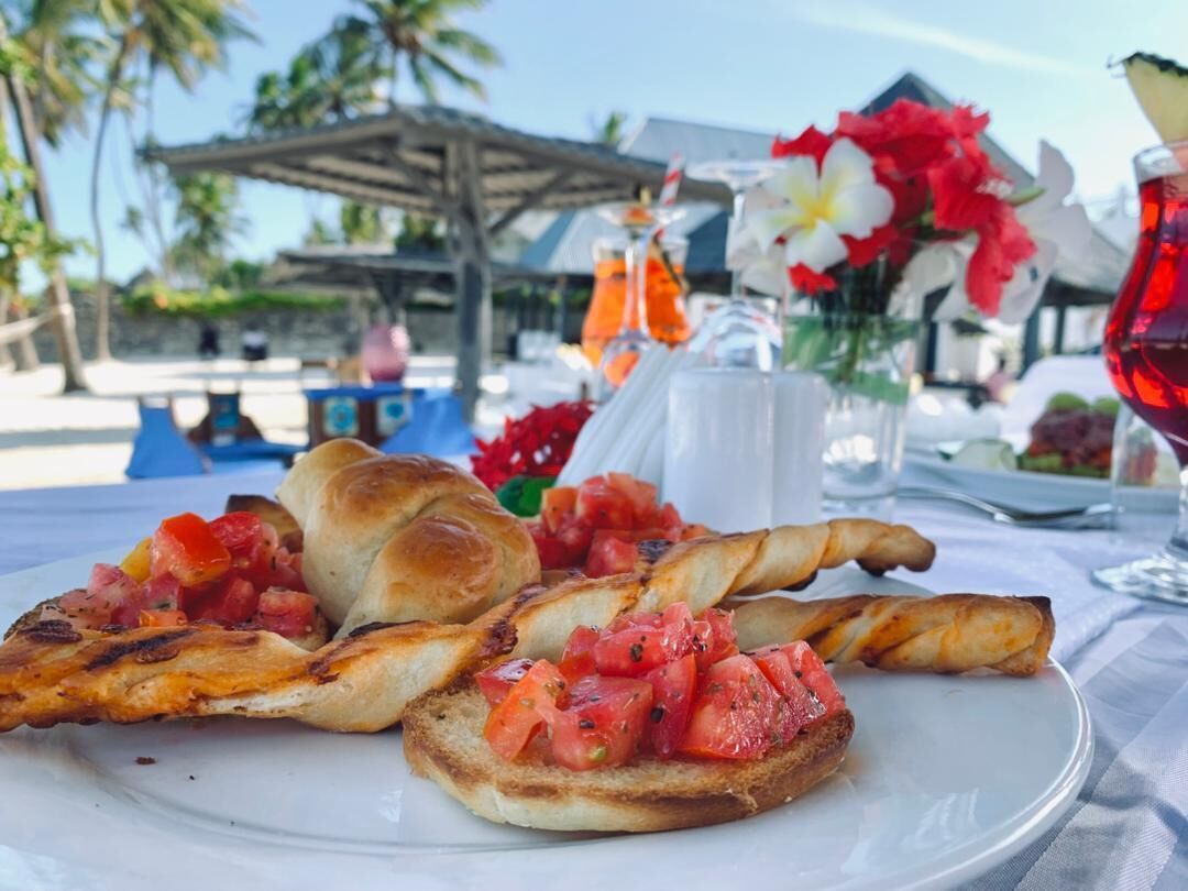 A plate with grilled chicken and bread, a glass of red drink, and a tropical setting with palm trees and a beach in the background.