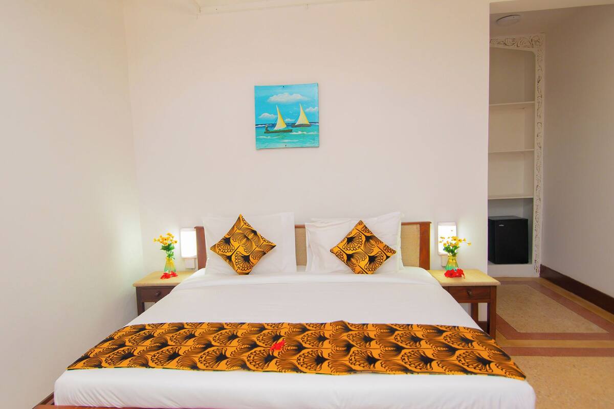 Una habitación de hotel con cama, mesita de noche, una pequeña mesa con un florero y un cuadro en la pared.