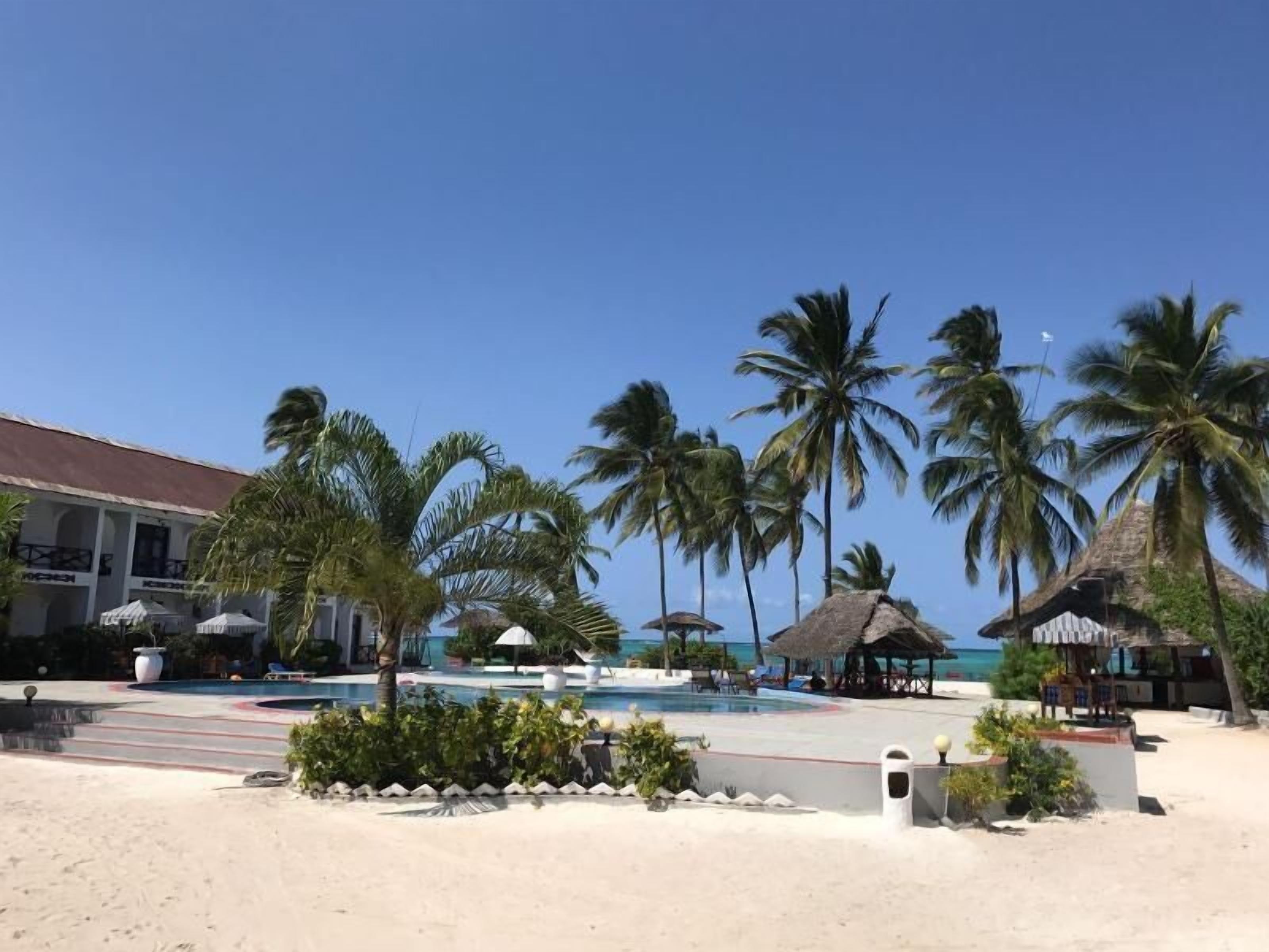 A beach resort with a pool, palm trees, and a building with a thatched roof.