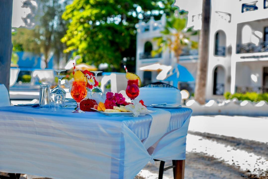 A table set outdoors with a blue tablecloth, clear glasses, and a variety of fruits and flowers.