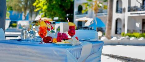 A table set outdoors with a blue tablecloth, clear glasses, and a variety of fruits and flowers.