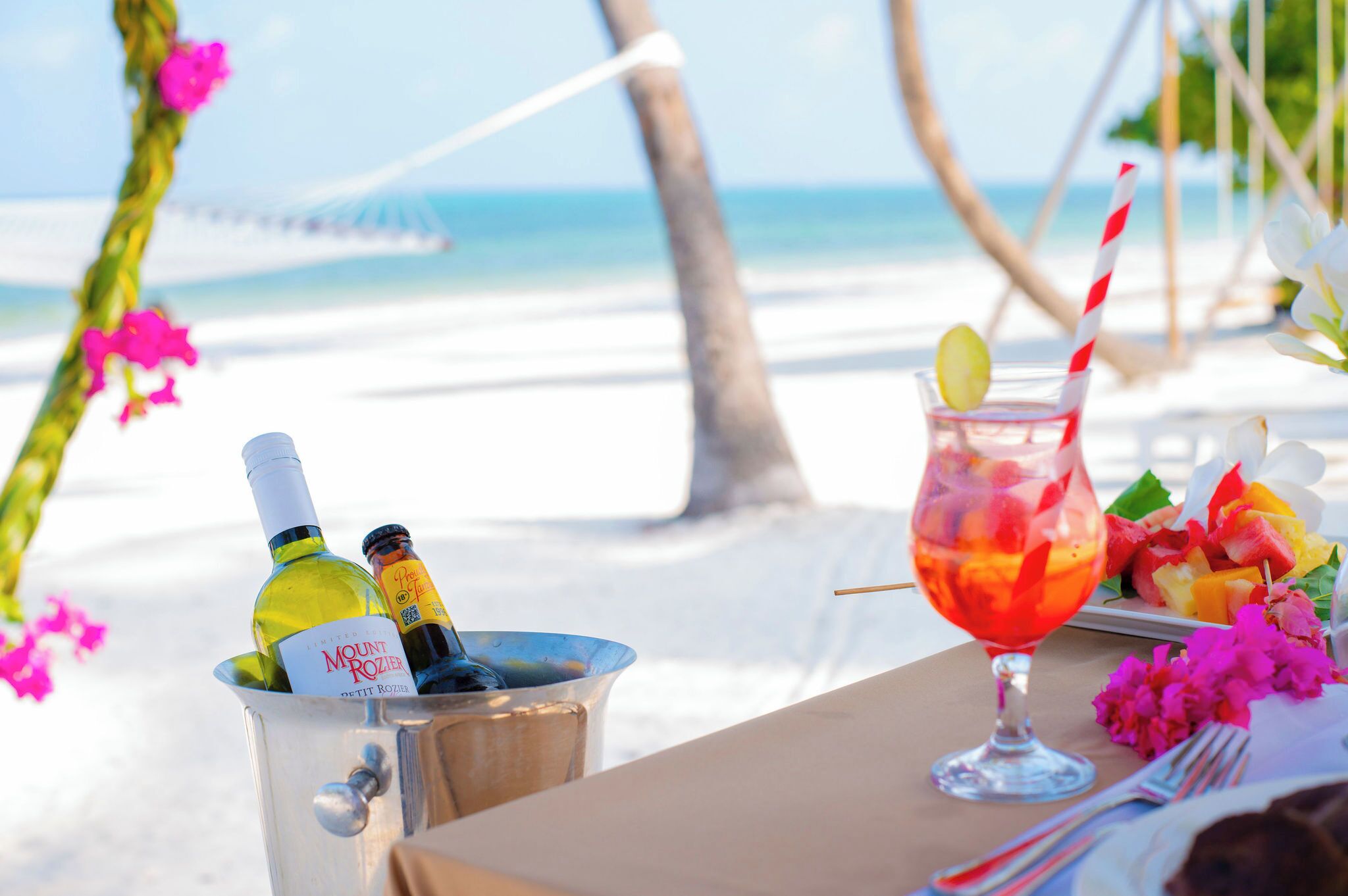 Una mesa frente al mar, con una bebida, una botella de vino y un plato de fruta.