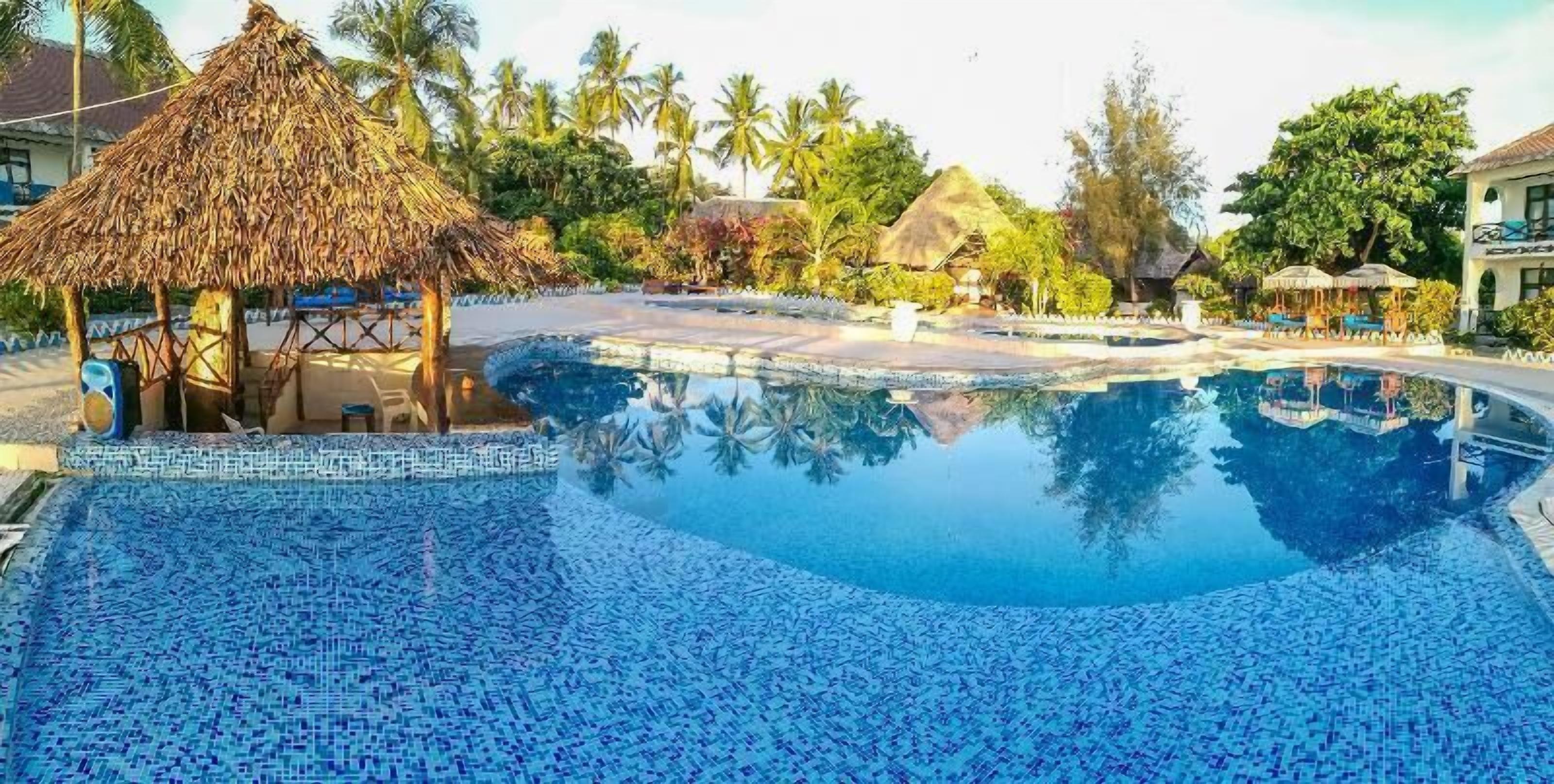 A swimming pool with mosaic tiles, surrounded by palm trees and a thatched-roof structure.