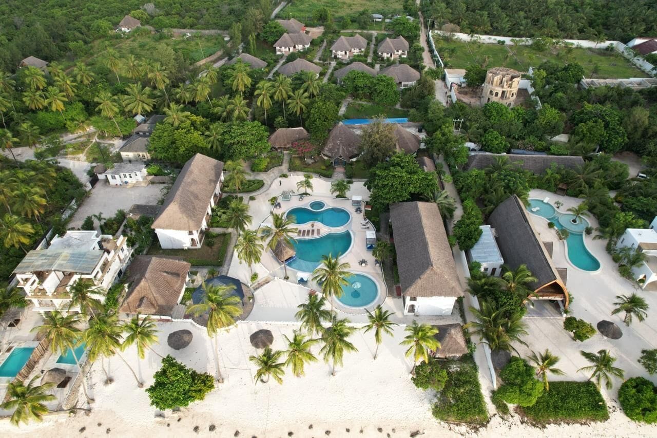 An aerial view of a resort with multiple buildings, palm trees, and a central swimming area with two pools.