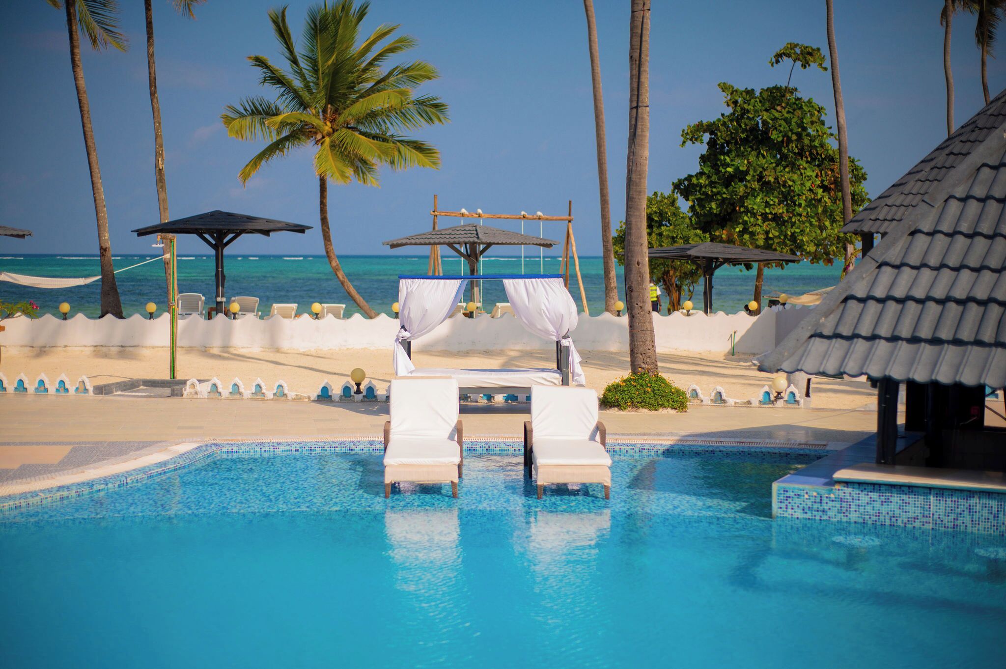A pool area with lounge chairs and a canopy, surrounded by palm trees and a view of the ocean.