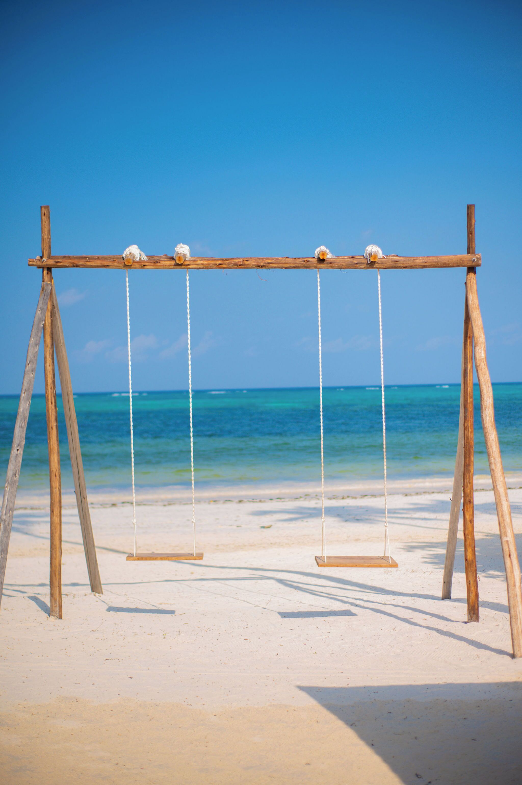 A wooden swing set on a sandy beach with clear blue water in the background.