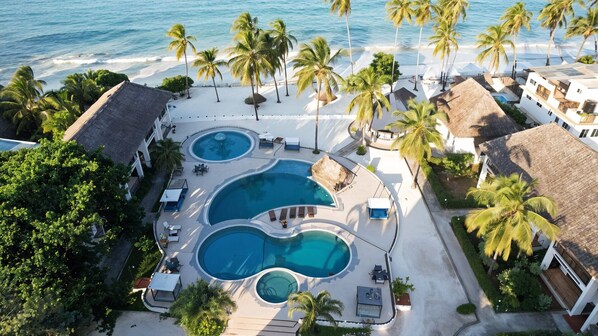 Vue aérienne d’un complexe hôtelier avec deux piscines, des palmiers et une plage.