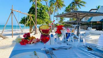 Un montaje para comer frente al mar con una mesa dispuesta para dos personas, que incluye una bebida tropical, una bandeja de frutas y flores.