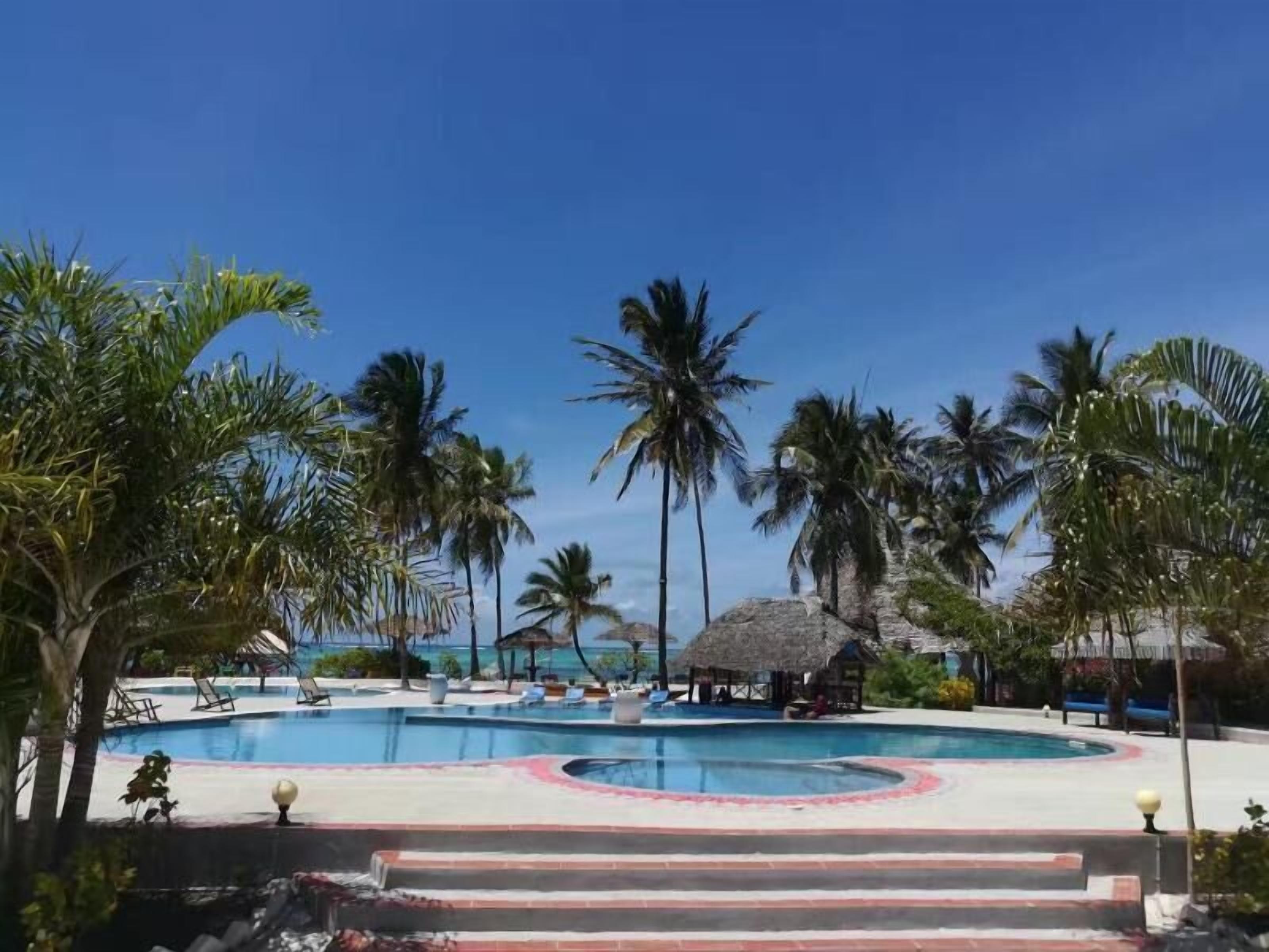A pool area with palm trees, lounge chairs, and a thatched-roof structure.