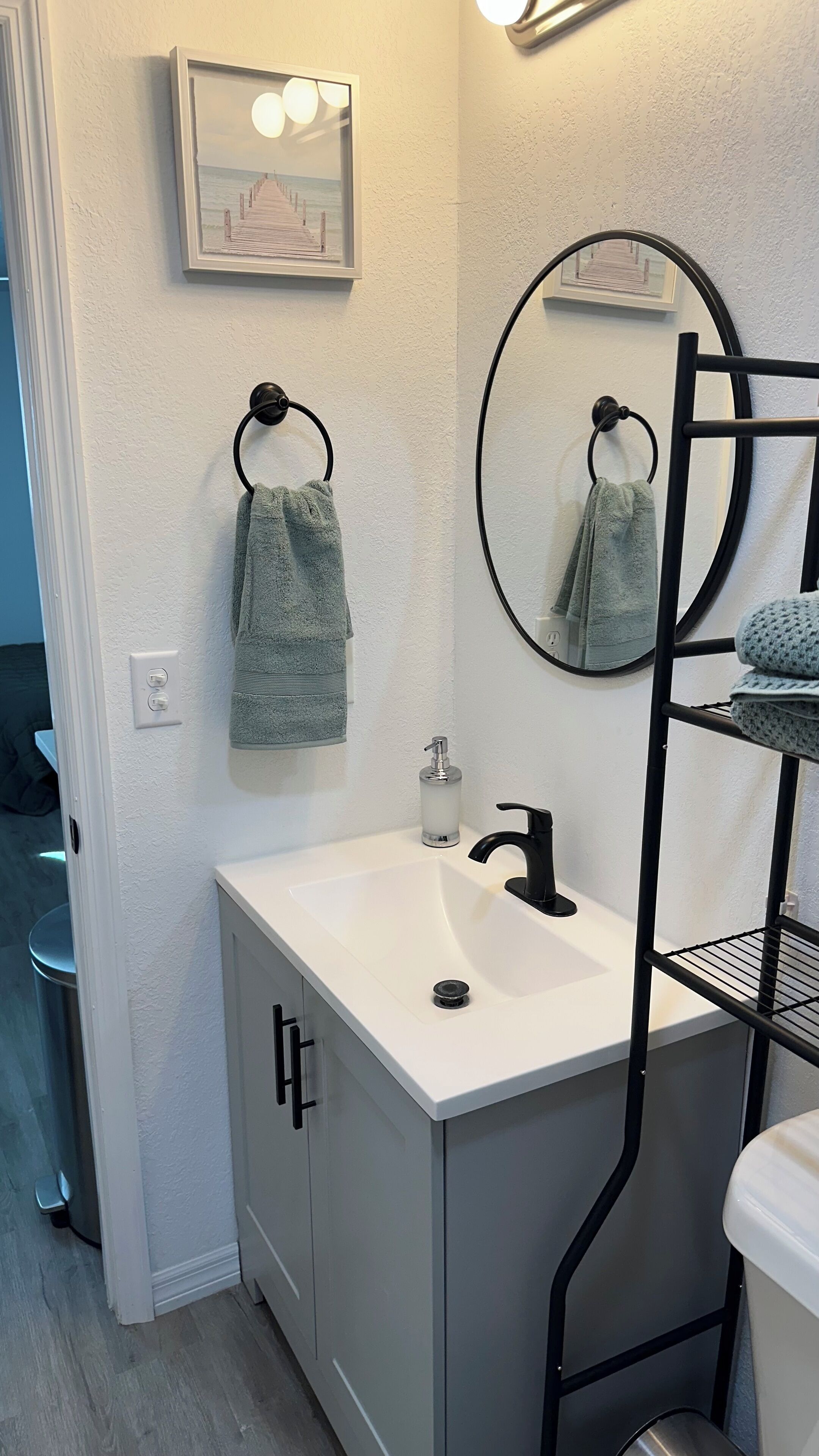 Une salle de bain avec un lavabo blanc, un porte-serviettes noir, un miroir et un tableau représentant une jetée accroché au mur.