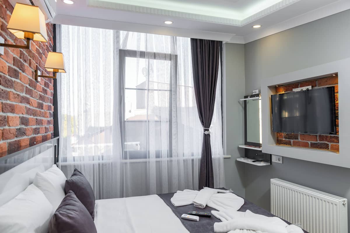 A hotel room with a brick wall, a large window with sheer curtains, a flat-screen TV mounted on a brick wall, a bed with white linens, and a bedside table with a remote control.