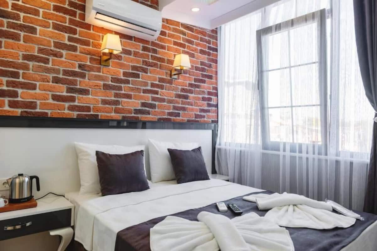 A hotel room with a brick wall, a bed with white and dark pillows, a bedside table with a kettle, and a window with sheer curtains.