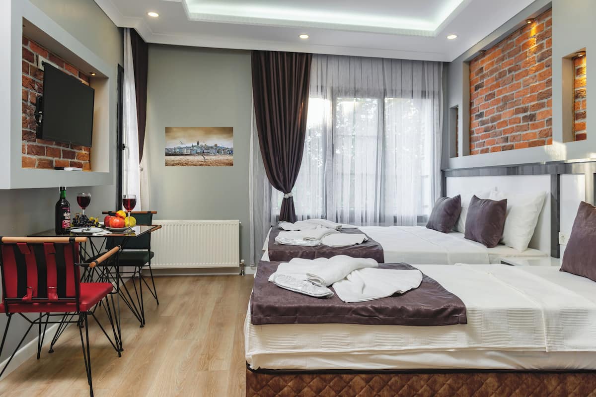 A hotel room with two beds, a dining area with a table and chairs, a TV mounted on a brick wall, and a window with curtains.