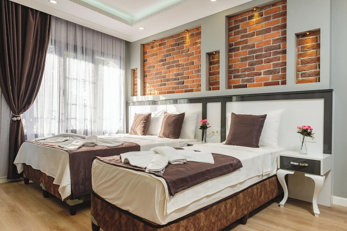 A hotel room with two beds, a brick accent wall, a nightstand with a vase of flowers, and a large window with curtains.
