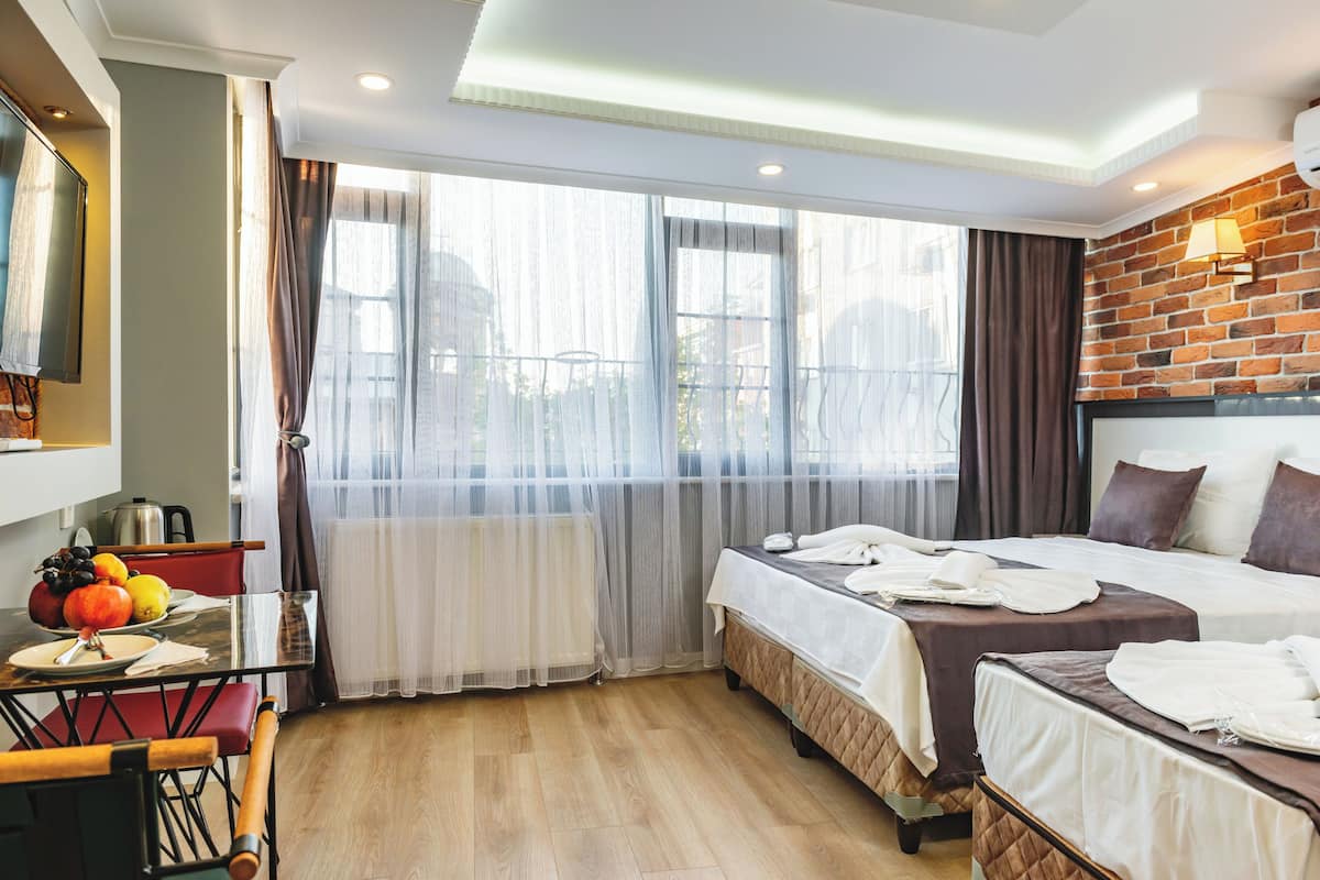 A hotel room with two beds, a dining table with fruit, a kitchen area, and large windows with sheer curtains.