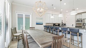 A dining area with a wooden table and chairs, a kitchen with white cabinets and bar stools, and pendant lighting. - Coastal Dream Home | Ocean Views & Rooftop Spa (Hilton Head Island)