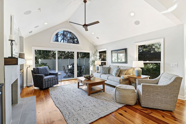 A spacious living room with a fireplace, a ceiling fan, and a large window.