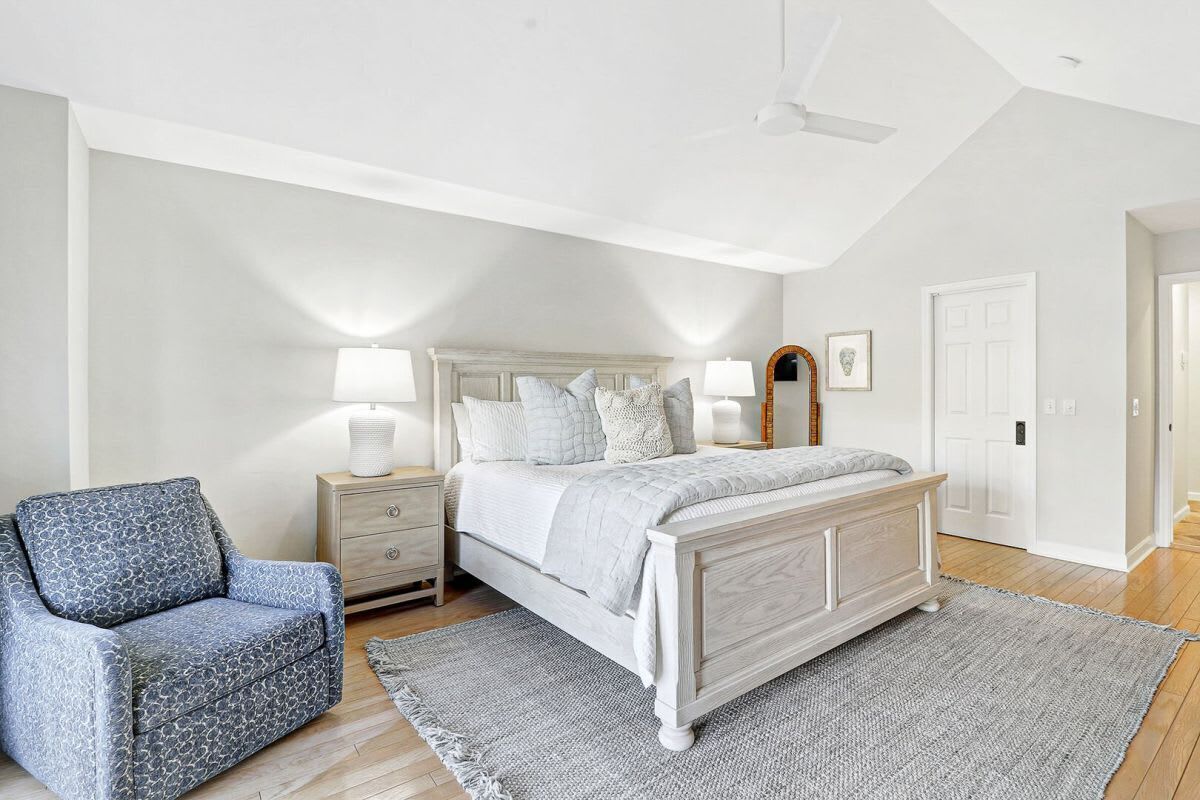 A bedroom with a bed, a chair, a nightstand, a lamp, a ceiling fan, and a door.