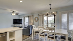 A dining area with a table and chairs, a ceiling fan, a television, and a window with blinds. - Coastal Getaway | Ocean Views & Resort Amenities (Hilton Head Island)