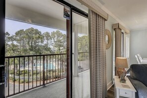 A balcony with a view of a swimming pool and trees, a sofa, a lamp, and a small table.