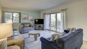 Una sala de estar con sofá, sillón, mesa de centro, televisor y ventanas amplias.