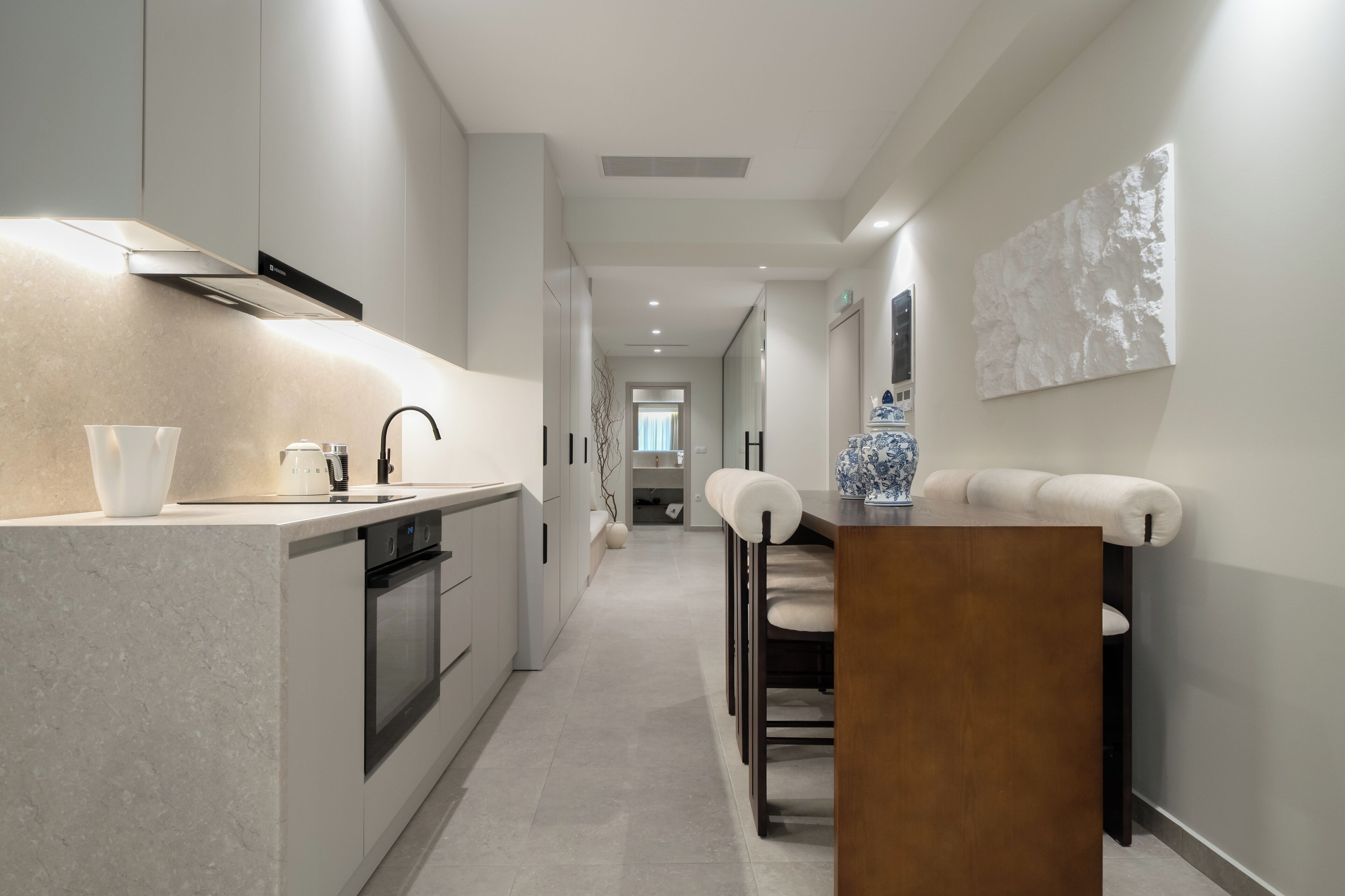 A modern kitchen with a central island, a dining area with a wooden table and chairs, and a hallway leading to a bathroom.