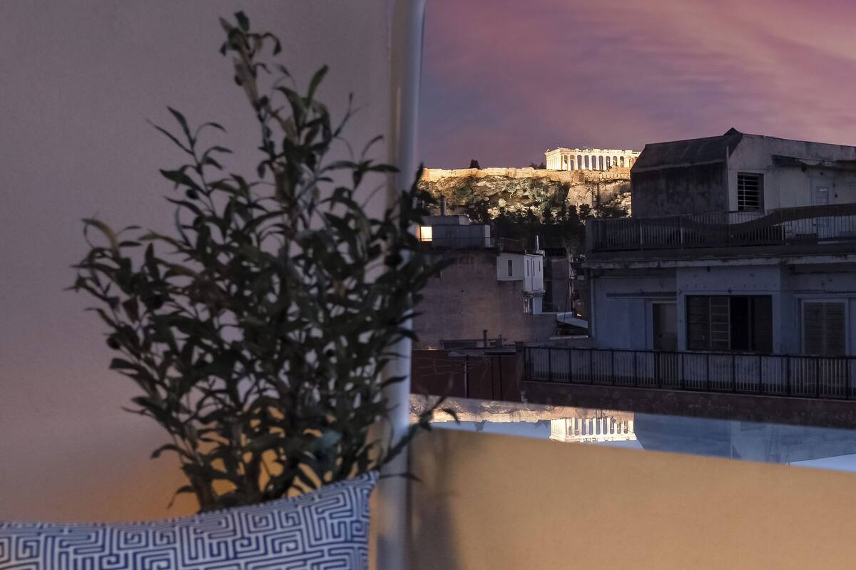 A balcony with two wine glasses, a potted plant, and a view of buildings against a twilight sky.