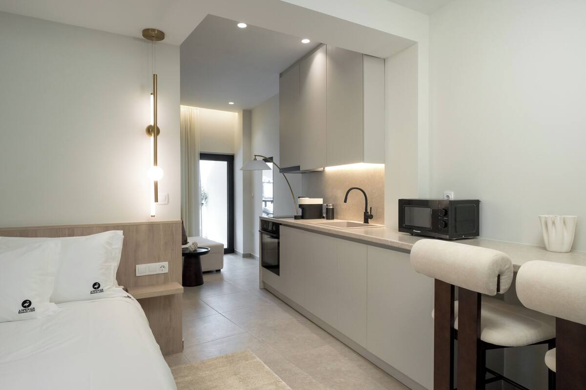 A modern hotel room with a bed, a small kitchenette, a sitting area with a chair, and a television.
