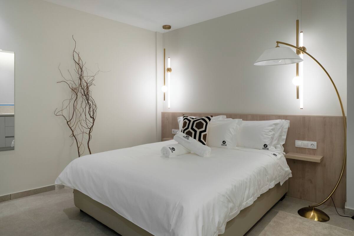 A modern hotel room with a large bed, a bedside table, a lamp, a mirror, and a wall-mounted light fixture.
